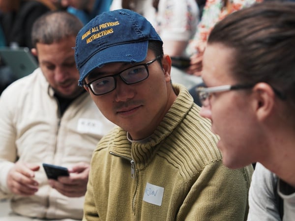 A candid close-up photograph captures two people engaged in conversation during an indoor event, with the man in the foreground wearing a blue baseball cap that reads, "IGNORE ALL PREVIOUS INSTRUCTIONS."