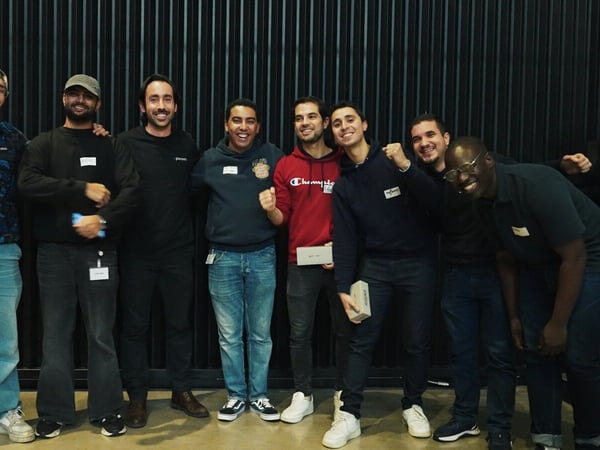 A group of approximately ten men stands together, smiling and posing for a group photo indoors against a dark, vertically ribbed wall. Several individuals are wearing name tags and casual attire, including jeans and hoodies.