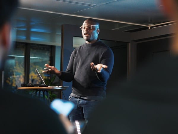A man wearing glasses and a dark sweater is giving a presentation or talk in a dimly lit, modern indoor setting, captured over the blurred shoulders of two audience members.