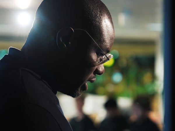 A dramatic, low-key photograph captures the profile of a bald Black man wearing glasses, heavily backlit and silhouetted against a blurred background of colorful bokeh lights, suggesting he is speaking or focused intently at an indoor event.