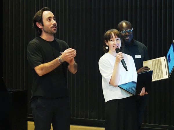 A candid photograph taken indoors showing three people on a stage or presentation area, where a woman in the center is speaking into a microphone while holding a laptop.