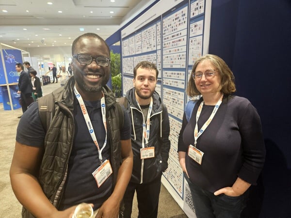 A group of three conference attendees, two men and one woman, stand side-by-side smiling at the camera in front of a large, detailed technical poster board at an indoor exhibition hall.