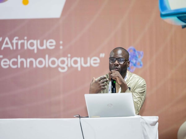 A man wearing glasses and a traditional gold-colored garment is speaking into a microphone at a podium with a laptop. He is positioned in front of a large banner featuring French text about technology in Africa.
