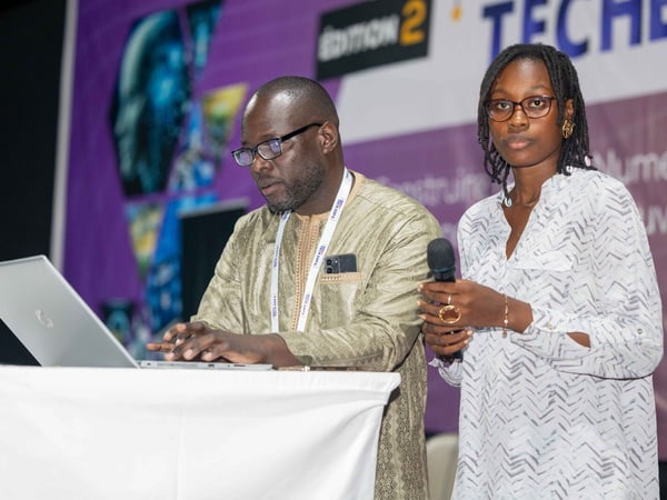 A man and a woman are at a technology event; the man is typing on a laptop at a podium while the woman stands beside him holding a microphone.