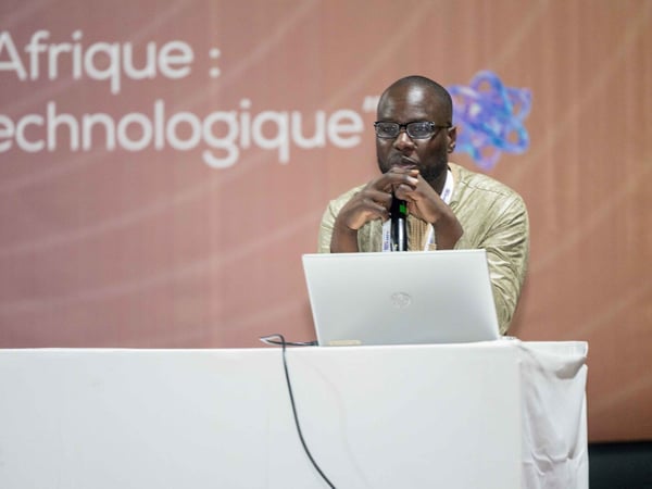 A man wearing glasses and a traditional gold-colored garment sits at a table with a laptop, speaking into a microphone during a tech event.