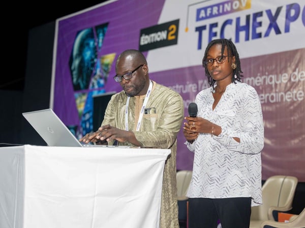 A man and a woman are on a stage during a tech event; the man is typing on a laptop while the woman stands beside him holding a microphone.
