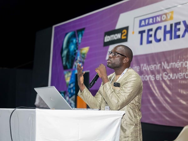 A man in traditional attire is speaking into a microphone while standing behind a podium with a laptop at a tech event.
