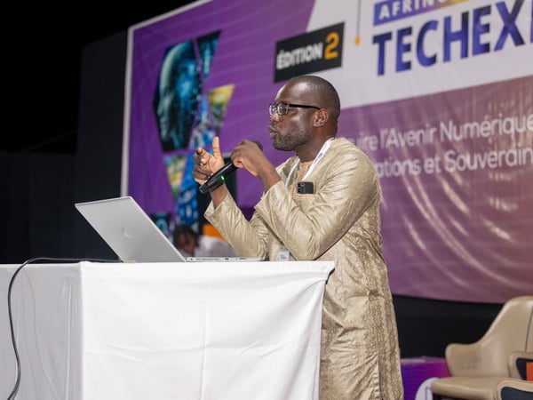 A man in traditional gold-colored attire is giving a presentation at a podium with a laptop and a microphone. The background features a large purple banner for the Afrinov Tech Expo.