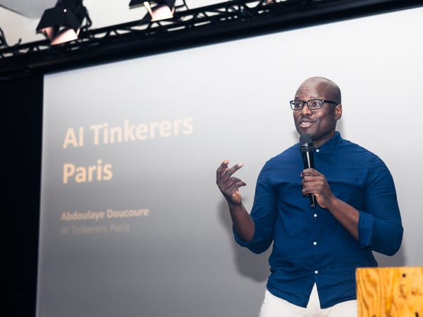 A man is giving a presentation on stage, holding a microphone in front of a screen that reads 'AI Tinkerers Paris'.