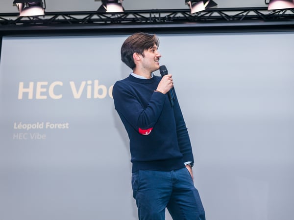 A man stands on a stage holding a microphone, giving a presentation in front of a screen that reads 'HEC Vibe'.