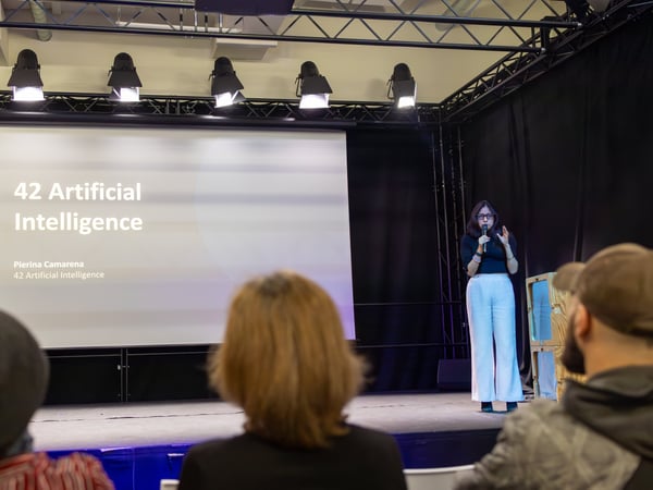 A woman stands on a stage giving a presentation about '42 Artificial Intelligence' to an audience.