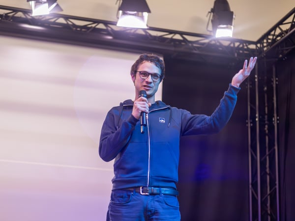 A man wearing glasses and a blue hoodie is giving a presentation on a stage, holding a microphone and gesturing with his hand. He stands in front of a large screen under stage lighting.