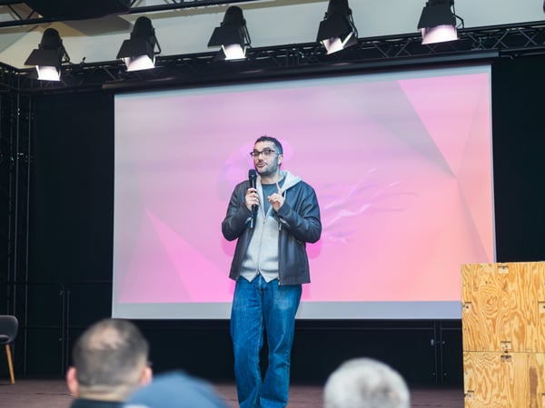 A man stands on a stage giving a presentation, holding a microphone in front of a large screen with a pink abstract background.