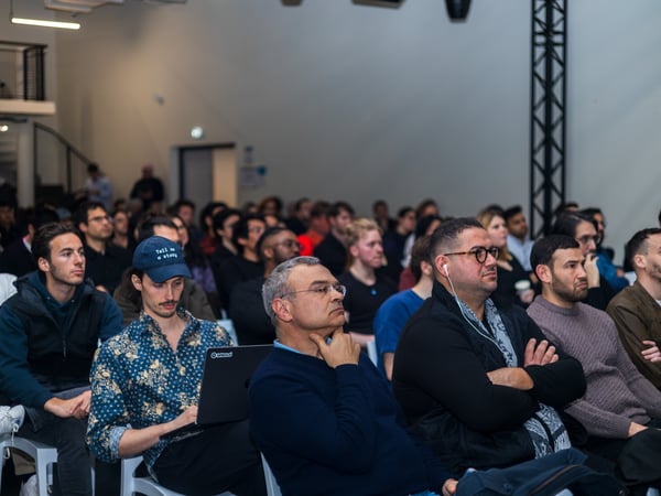 A large audience is seated in an indoor venue, attentively listening to a presentation or talk. Most individuals are looking toward the front of the room, with some using laptops.