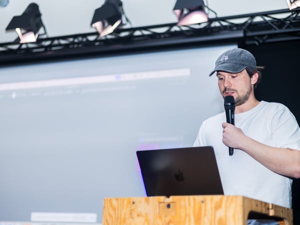 A man in a white t-shirt and grey baseball cap is giving a presentation, speaking into a microphone while looking at a laptop on a wooden podium.