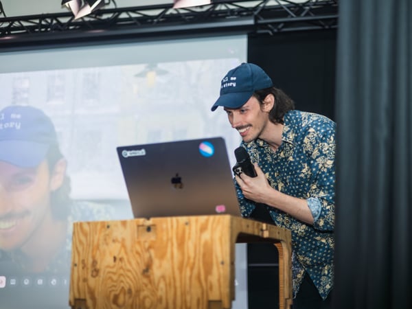 A man wearing a blue cap and a patterned shirt is smiling while looking at a laptop on a wooden podium during a presentation.