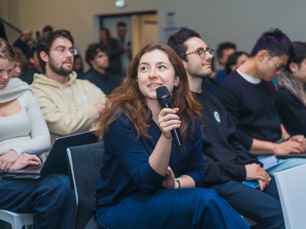 A young woman sitting in an audience holds a microphone, looking attentively towards a speaker off-camera during a Q&A session.