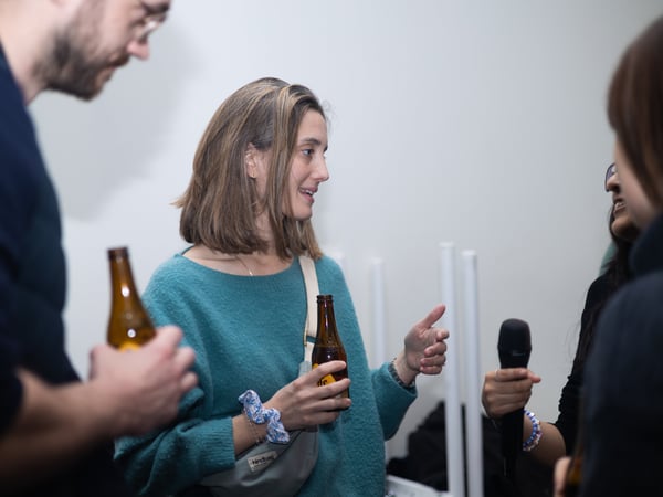 A group of people are socializing in an indoor setting, with a woman in a teal sweater holding a beer bottle while engaged in conversation.