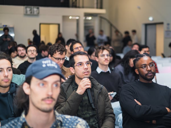 A photograph of an audience at an indoor event, featuring a man in the center holding a microphone and speaking while others listen attentively.