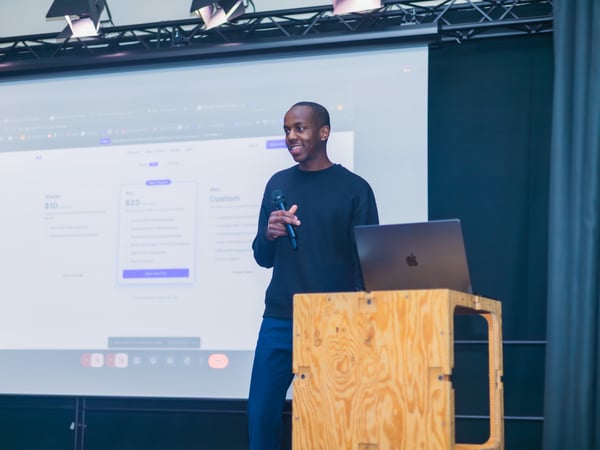 A man stands on a stage giving a presentation, holding a microphone next to a wooden podium with a laptop, with a large screen behind him displaying pricing plans.