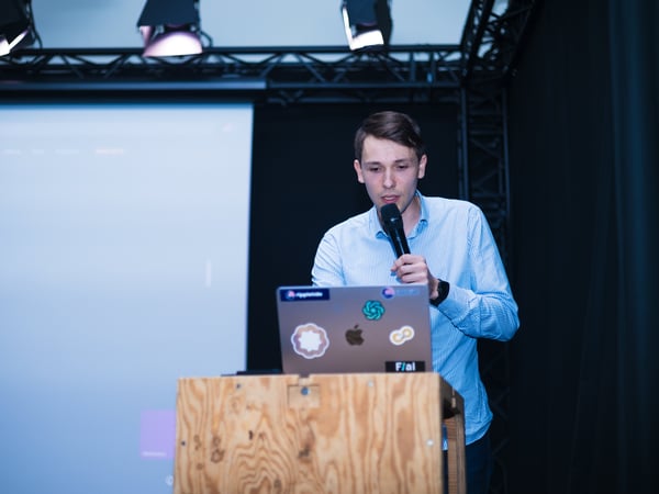 A man in a light blue shirt stands behind a wooden podium, speaking into a microphone while giving a presentation with a laptop and projector screen.