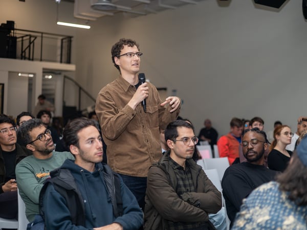 A man in a brown corduroy shirt stands in an audience and speaks into a microphone during a presentation or event.