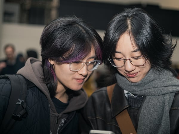 Two young women with glasses are looking down at a mobile phone together in a crowded indoor setting.