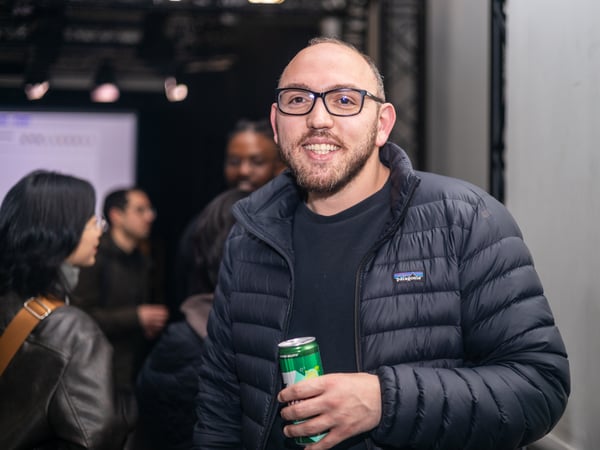 A man with glasses and a beard smiles at the camera while holding a green beverage can in a social indoor setting.
