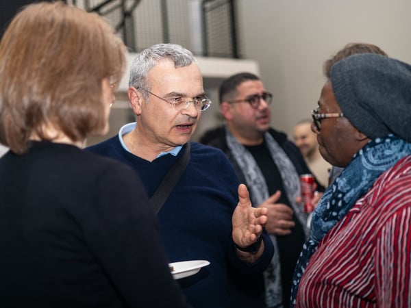 A group of people are engaged in a conversation at an indoor social event, with a man in glasses gesturing as he speaks.