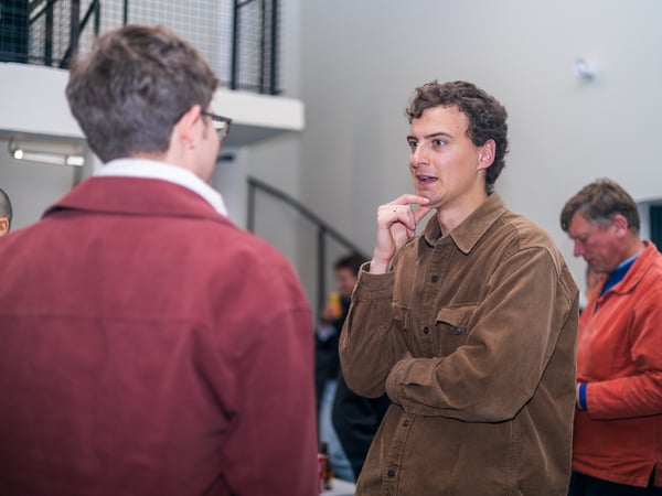 A man in a brown corduroy shirt is engaged in a conversation with another person at an indoor social gathering.