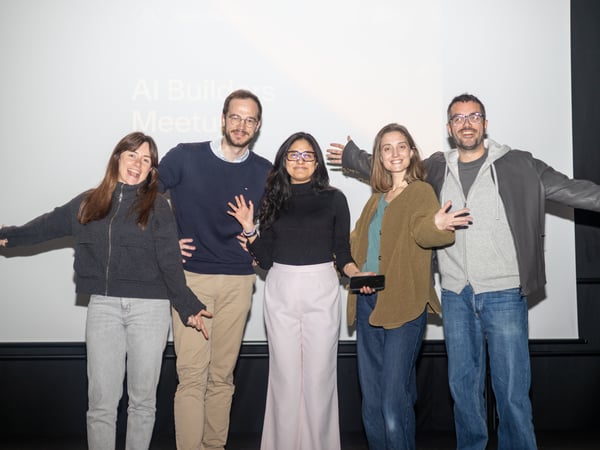A group of five smiling people posing for a photo in front of a projection screen that reads 'AI Builders Meetup'.