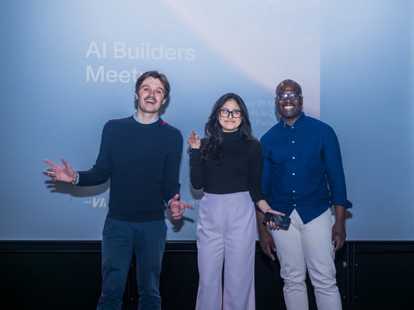 Three people, two men and one woman, are smiling and posing for a photo in front of a projection screen for an AI Builders Meetup event.