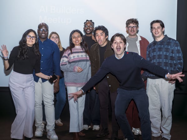 A group of nine people posing playfully for a photo in front of a projection screen that reads 'AI Builders Meetup'.