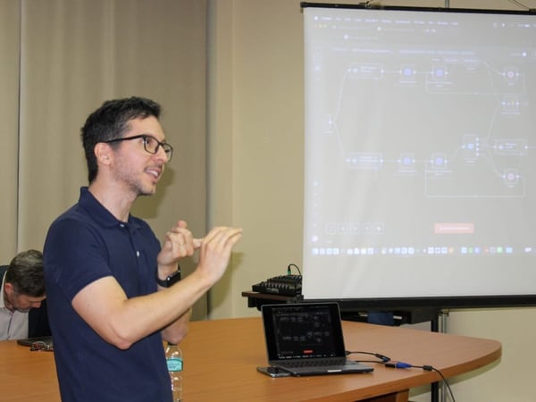 A man wearing a blue polo shirt and glasses is giving a technical presentation, gesturing with his hands in front of a large projection screen displaying a complex workflow or data flow diagram.