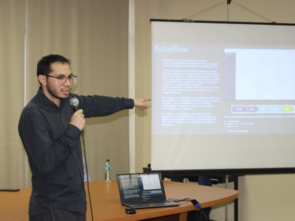 A man is giving a technical presentation indoors, holding a microphone and pointing toward a screen displaying slides about Kubeflow.