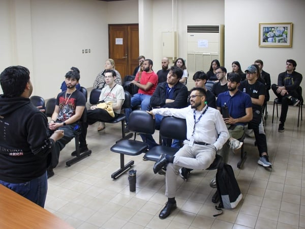 A candid photograph captures a speaker, seen from the back, addressing a seated audience of approximately 17 young adults in a brightly lit indoor meeting or classroom setting.