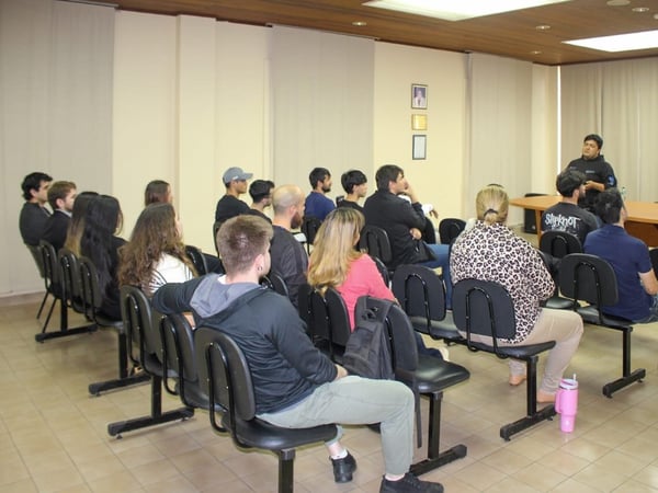 A candid photo taken inside a lecture hall or conference room, showing an audience seated in rows of black chairs facing a male speaker who is standing behind a long wooden presentation table.