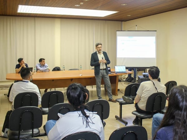 A man in a blazer stands presenting with a microphone in a conference room, addressing an audience seated in the foreground, while two other men are seated behind a long panel table.