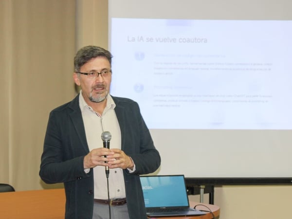 A man wearing glasses and a dark jacket stands indoors holding a microphone, giving a presentation in front of a large projection screen displaying Spanish text about AI becoming a co-author.