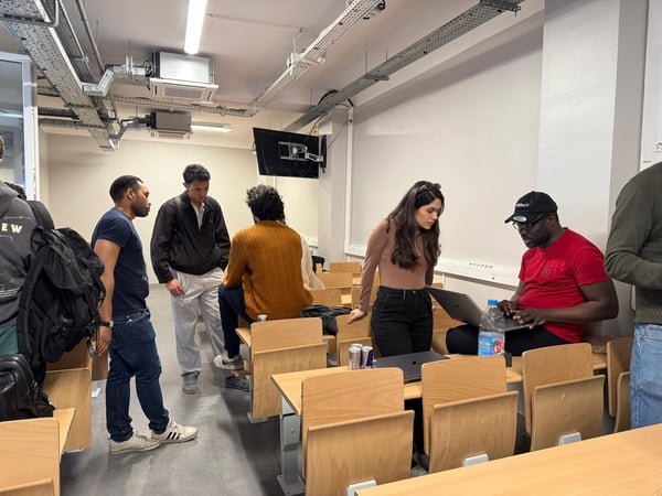 A group of young adults are gathered in a classroom or lecture hall setting, with some standing in conversation and others sitting at desks with laptops and drinks.