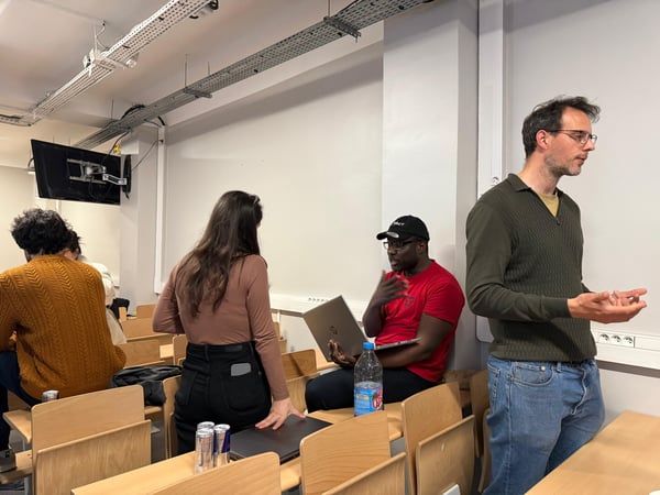 A group of people are gathered in a lecture hall or classroom setting, with one man standing and gesturing while others sit with laptops.