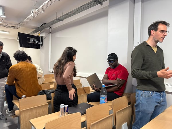A group of people are gathered in a classroom or lecture hall setting, with some sitting at wooden desks and others standing and talking.