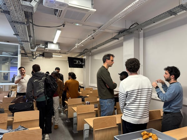 A group of people are gathered in a lecture hall or classroom, some standing and talking while others sit, with a tray of croissants visible in the foreground.