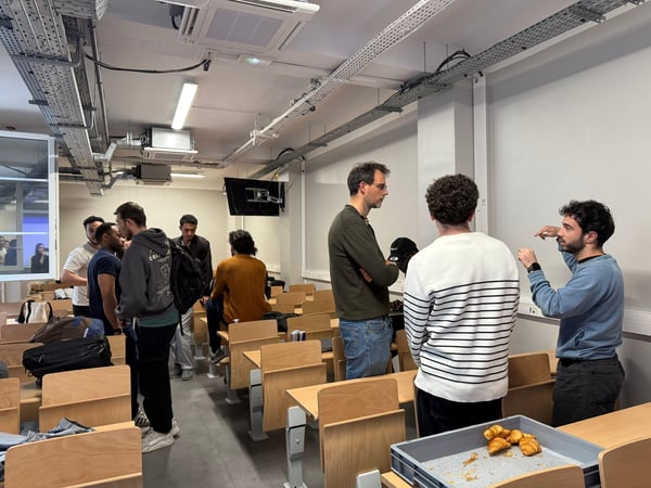 A group of people are gathered in a classroom or lecture hall, some standing and talking while others are seated, with a tray of croissants in the foreground.