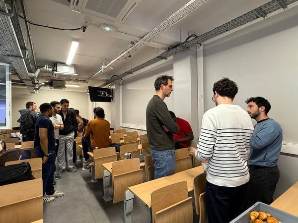 A group of young men are gathered in a lecture hall, standing and sitting among rows of wooden desks while engaged in conversation.