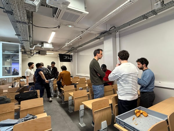 A group of young men are gathered in a modern classroom or lecture hall with exposed ceiling pipes, socializing in small groups near rows of wooden desks.