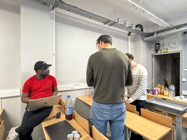 A group of men are in a classroom or office setting; one person sits on a desk with a laptop while others stand near a table with drinks and snacks.