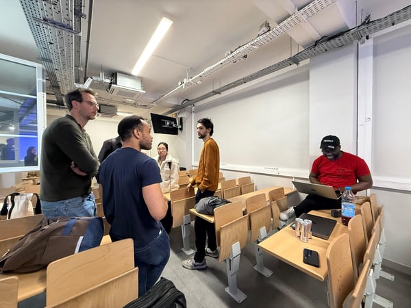 A group of young adults are gathered in a modern classroom or lecture hall, with some standing in discussion while one person sits working on a laptop.