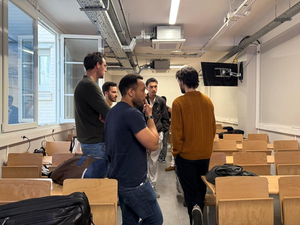A group of men are engaged in a discussion inside a classroom or lecture hall featuring rows of wooden desks and industrial ceiling fixtures.
