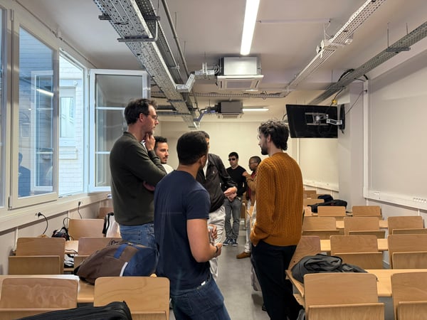 A group of men are standing and talking in a classroom or lecture hall featuring rows of wooden desks and exposed ceiling conduits.